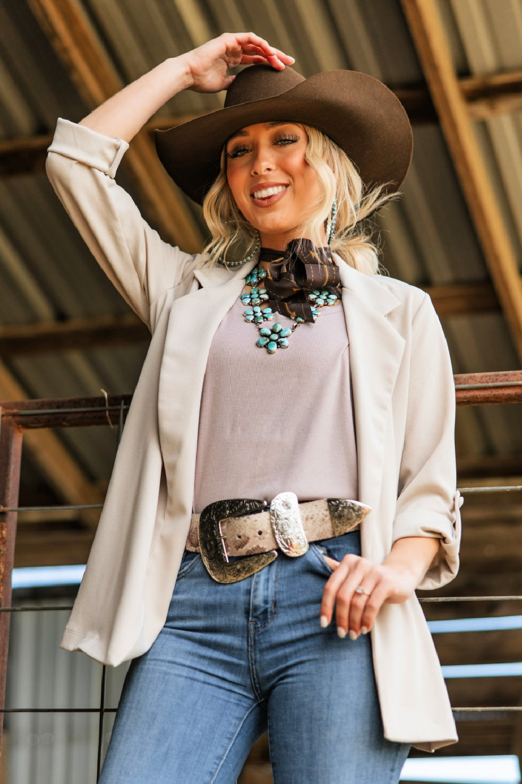 A woman wearing a neutral ivory color boyfriend blazer with a cowgirl hat, and standing in a barn setting.