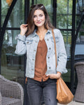 Woman wearing a light wash denim jacket, brown shirt, and carrying a tan bag outdoors.