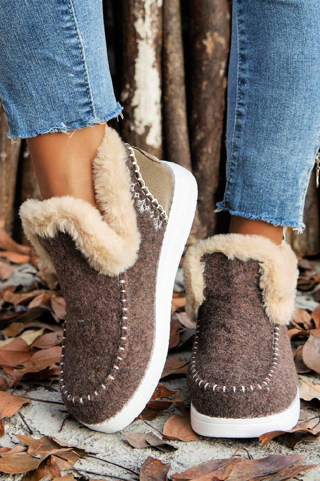 Brown slip-on slippers with fur lining worn by a person on a leaf-covered ground.