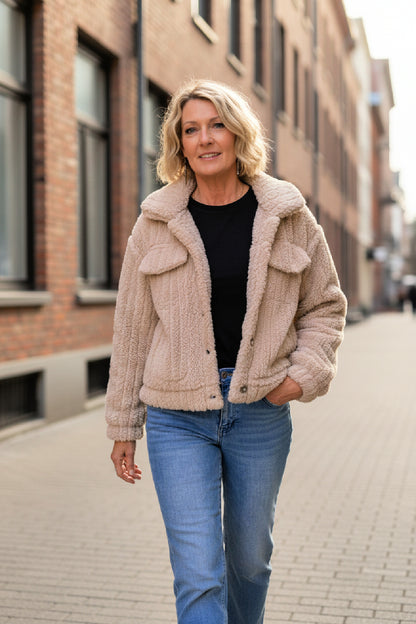 Woman wearing a beige furry cropped jacket and blue jeans walking down a street.