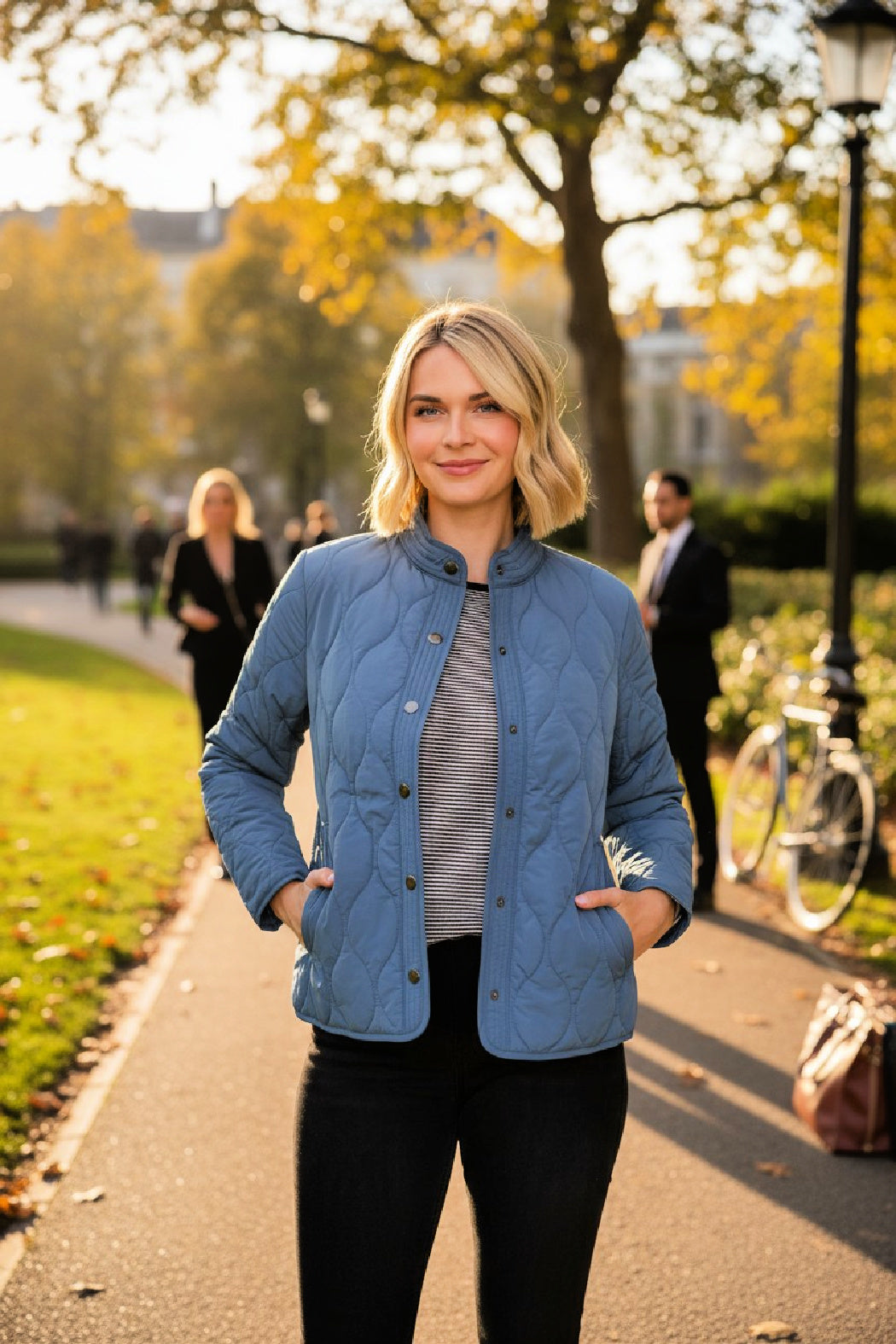 Woman wearing a blue lightweight quilted barn jacket in an outdoor city park setting with trees and people in the background.