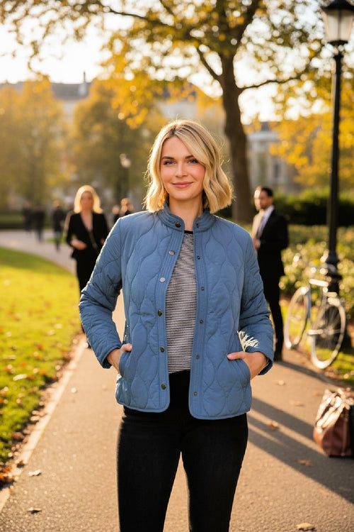 Woman wearing a blue lightweight quilted barn jacket in an outdoor city park setting with trees and people in the background.